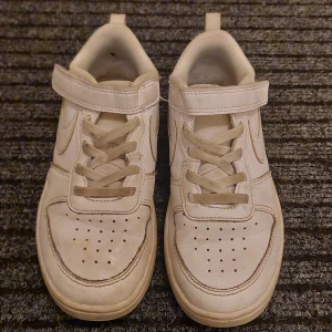 Vita sneakers från Nike - Säljer ett par vita Nike sneakers i storlek EUR 33. De har snörning och kardborreband för extra säkerhet. Skorna är i syntetmaterial och har en klassisk Nike-logga på sidan. Perfekta för vardagsbruk och lek. De är välanvända men fortfarande i bra skick.