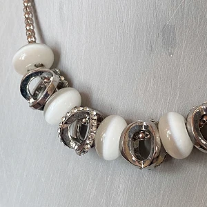 Elegant halsband med vita och silverfärgade detaljer - Snyggt halsband med vita pärlor och silverfärgade detaljer. Perfekt för att ge en touch av elegans till din outfit. Kedjan är i en fin flätad stil som kompletterar de glänsande detaljerna. Passar både till vardags och festliga tillfällen.