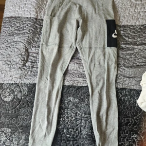Grå mjukisbyxor från Nike - Säljer ett par grå tights från Nike i storlek S. Tightsen har en bekväm passform och är perfekta för träning eller chill. De har en svart ficka med Nike-loggan på sidan, vilket ger en cool detalj samt ficka på andra låret också. 