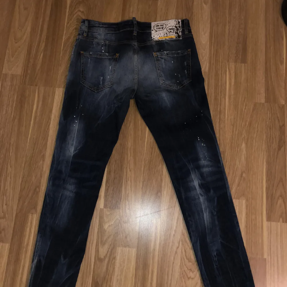 Säljer ett par snygga mörkblå jeans från Dsquared2. De har en cool tvättad look med ljusare partier och små färgstänk som ger dem en unik stil. Jeansen har en klassisk femficksdesign och en knappgylf. Märkesdetaljer finns både på den röda fliken vid fickan och på den grafiska lappen bak. . Jeans & Byxor.