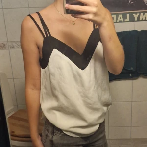 Vit och svart linne med dubbla axelband - Säljer ett snyggt linne i vitt med svarta detaljer. Det har en V-ringad design och dubbla axelband som ger en unik look. Perfekt för sommardagar eller en utekväll. Passar bra till jeans eller kjol! Den har två lager av vitt tyg som göra att den inte är genomskinlig. Köparen står för frakt