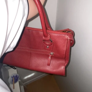 Röd handväska i skinn - Snygg röd handväska i skinn med dragkedja framtill. Perfekt för både vardag och fest. Väskan har en stilren design med två handtag och en praktisk storlek för alla dina nödvändigheter.