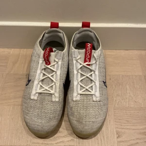 Nike Vapormax  - Säljer ett par gråa Nike Vapormax  med röda och vita detaljer. Skorna har en unik transparent sula och är i nyskick. Perfekta för både träning och vardagsbruk. De har en snygg snörning och är superbekväma att ha på sig. Skorna är aldrig använda, nypris ligger på 2000kr, mitt pris 1100kr