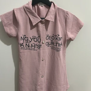 Rosa kortärmad skjorta från NINI Kids - Säljer en söt rosa kortärmad skjorta från NINI Kids. Skjortan har knappar framtill och ett tryck med text på bröstet. Perfekt för en avslappnad och lekfull stil. Passar bra till både vardag och festliga tillfällen.