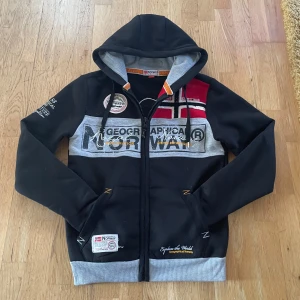 Svart och grå hoodie från Geographical Norway - Säljer en snygg svart och grå hoodie från Geographical Norway. Den har en dragkedja framtill och coola tryck både fram och bak. Perfekt för höst och vår, med en skön huva och långa ärmar.. Hör av dig vid intresse!