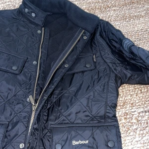 Svart quiltad jacka från Barbour - Säljer en snygg svart quiltad jacka från Barbour i storlek XL i ungdomsstorlek! Jackan har en stilren design med dragkedja och knappar framtill, samt flera praktiska fickor. Perfekt för höst och vår när du vill hålla stilen. 🖤