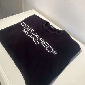 Svart sweatshirt från Dsquared2 - Säljer en snygg svart sweatshirt från Dsquared2 med vit text 'DSQUARED2 MILANO' på bröstet. Tröjan är i mycket bra skick och perfekt för både vardag och fest. Den har en klassisk passform och är superbekväm. Passar perfekt till jeans eller chinos!