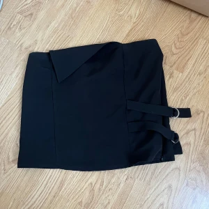 Svart topp med spännen - Snygg svart topp med två dekorativa spännen på sidan. Perfekt för en utekväll eller fest! Toppen har en stilren design och är lätt att matcha med olika underdelar. Den är i bra skick och redo för nya äventyr.