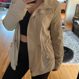 Beige jacka från ONLY - Säljer en snygg beige jacka från ONLY i storlek M. Jackan har en dragkedja och knappar framtill, samt en praktisk huva. Perfekt för höst och vår med sin stilrena design och bekväma passform. Den är i mycket bra skick och redo för nya äventyr!