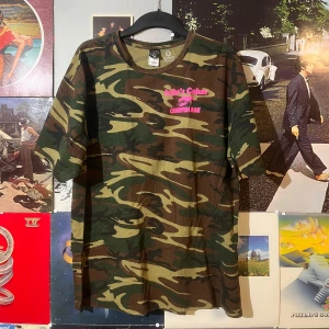 Vintage Camouflage t-shirt från JuJu's Cajun - Säljer en cool camouflage t-shirt från JuJu's Cajun med rosa tryck på bröstet och ryggen. T-shirten är i bomull och har en normal passform. Perfekt för en avslappnad stil eller för att sticka ut lite extra. Den är i mycket bra skick och redo för nya äventyr! Dm vid frågor