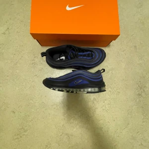 Nike Air Max 97 i svart och blå - Säljer ett par Nike Air Max 97 i svart och blått. De har en snygg vågig design med synlig Air-dämpning i sulan. Skorna är i mycket bra skick och kommer med originalkartong. Perfekta för både vardag och träning!