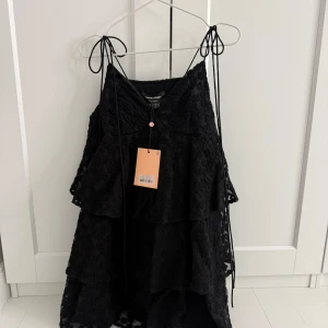 Svart spetsklänning från Custommade - Säljer en elegant svart spetsklänning från Custommade i storlek XS. Klänningen har ett vackert lager-på-lager utseende med tunna axelband och volangdetaljer. Perfekt för festliga tillfällen! Helt ny med tag kvar.