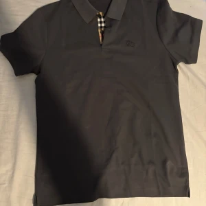 Svart pikétröja med rutiga detaljer - Säljer en stilren grå burberry pikétröja med klassisk krage och korta ärmar. Den har en snygg detalj med rutigt mönster vid knäppningen. Perfekt för både vardag och fest! 🖤