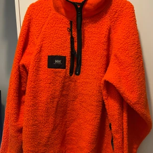 Orange fleece från Helly Hansen - Säljer min pappas Helly Hansen fleece. Tröjan har en dragkedja framtill och en liten logga på bröstet. Varm och perfekt för höst och vinter. Knappt använd bara ca 3 gånger, inga defekter.