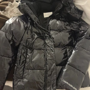 Svart pufferjacka från Calvin Klein Jeans - Säljer en snygg svart pufferjacka från Calvin Klein Jeans. Jackan har en glansig finish och är perfekt för kalla dagar. Den har en dragkedja framtill och en hög krage för extra värme. Perfekt för vintern och hösten! Pris kan diskuteras❤️köptes förra vintern har använts typ 2 gånger därför säljer vidare❤️