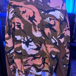 Camouflage hoodie från Adidas - Säljer en cool hoodie från Adidas med ett unikt camouflage-mönster i brunt, rosa och beige. Perfekt för höst och vår, den har en avslappnad passform och är superbekväm. Adidas-loggan syns diskret på bröstet. Perfekt för både chill och streetwear-stil!