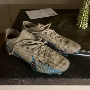 Nike fotbollsskor i vitt och blått - Säljer ett par Nike fotbollsskor i vitt med blå och röda detaljer. Skorna har snörning och är i bra skick, perfekta för fotbollsträning. De har en snygg design med Zoom-teknologi för extra komfort och prestanda på planen.