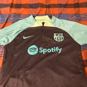 PLAYER EDITION FC Barcelona fotbollströja 23/24 - Säljer en FC Barcelona fotbollströja i mycket bra skick. Tröjan är brun med ljusblå ärmar och har klubbens emblem samt Nike-logga på bröstet. Den har en halv dragkedja vid halsen och är kortärmad. Perfekt för alla Barca-fans! 💙❤️byxa går att Få med 