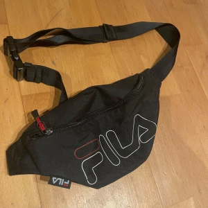 Svart magväska från Fila - Säljer en snygg svart magväska från Fila. Den har en justerbar rem och ett stort fack med dragkedja. Perfekt för att ha med sig det viktigaste på språng. Fila-loggan i vitt och rött ger en sportig touch. Använd den till vardags eller på festivaler!