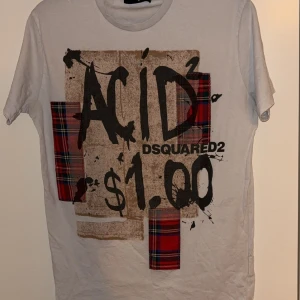 Vit t-shirt från Dsquared2 - Säljer en cool vit t-shirt från Dsquared2 i storlek XL.  Men sitter mer som L pga att den sitter lite slim. Den har ett unikt tryck med texten 'ACID $1.00' och röd-rutiga detaljer. Perfekt för en avslappnad stil eller fest. T-shirten är i bra skick och gjord av mjuk bomull. Tillverkad i Italien.