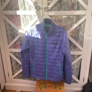 Lila pufferjacka från Patagonia - Säljer en snygg lila pufferjacka från Patagonia i storlek S. Jackan har en dragkedja och turkosa detaljer vid fickorna. Perfekt för kyliga dagar och har en modern look. Passar bra till både vardag och utflykter. 