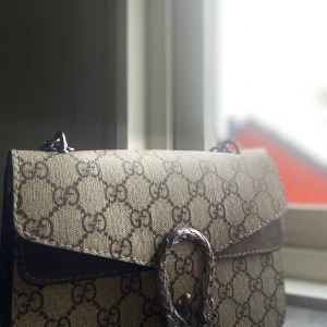 Mönstrad axelväska - Säljer en snygg axelväska med ett klassiskt mönster. Väskan har en elegant spänndetalj framtill och är perfekt för både vardag och fest. Den är i mycket bra skick och redo för en ny ägare!