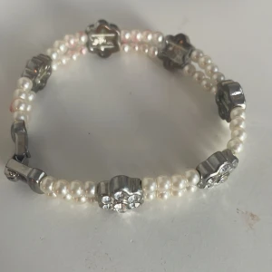Pärlarmband med blommiga detaljer - Snyggt armband med vita pärlor och blommiga detaljer i silverfärg. Perfekt för att ge en elegant touch till din outfit. En liten Sten fattas vid låset.. Armbandet har en elastisk passform vilket gör det enkelt att ta på och av. Passar både till vardags och festliga tillfällen. 🌸✨