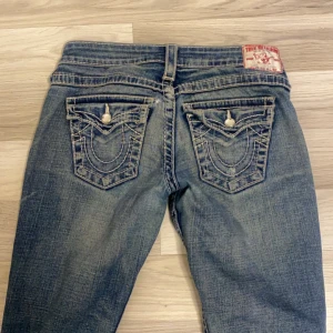 Blå jeans från True Religion - Säljer ett par snygga blå jeans från True Religion i bra skick. De har coola detaljerade bakfickor med knappar. Perfekta för en avslappnad stil!