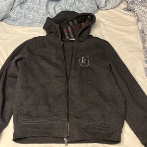 Grå huvtröja från Burberry - Säljer en snygg grå huvtröja från Burberry i mycket bra skick använd 3 gånger . Den har en dragkedja framtill och ett klassiskt rutigt mönster på insidan av huvan. Perfekt för höst och vår, med en skön passform och tillverkad i bomull och polyester. En stilren och bekväm tröja för vardagsbruk.Säljer den då den är för stor för mig och den är köpt på serevalle outlet i Italien. kvitto finnssss!!! Pris kan diskuteras vid snabb affär 