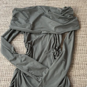 Oliv färgad tröja - Säljer en snygg grå offshoulder tröja i mjukt material. Perfekt för både vardag och fest! Tröjan har en smickrande passform med rynkdetaljer längs sidorna. Den är i mycket bra skick och redo för nya äventyr. Passar perfekt till höst och vår.