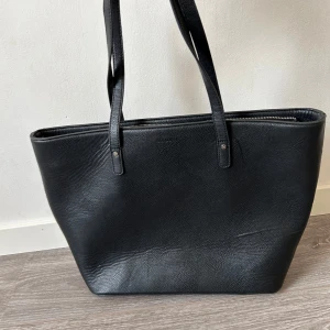 Svart axelväska i skinn - Snygg och rymlig svart axelväska i skinn med dragkedja upptill. Perfekt för vardagsbruk med en praktisk ytterficka med dragkedja. Insidan är ljus och har flera fack för enkel organisering. Väskan är i mycket bra skick och passar till alla säsonger.