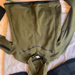 Olivgrön jacka från Nike - Säljer en olivgrön jacka från Nike i mycket bra skick. Den har en dragkedja framtill och är långärmad med en praktisk ficka på ärmen. Perfekt för höst och vår, tillverkad i ett mjukt syntetmaterial. Passar både till träning och vardagsbruk.