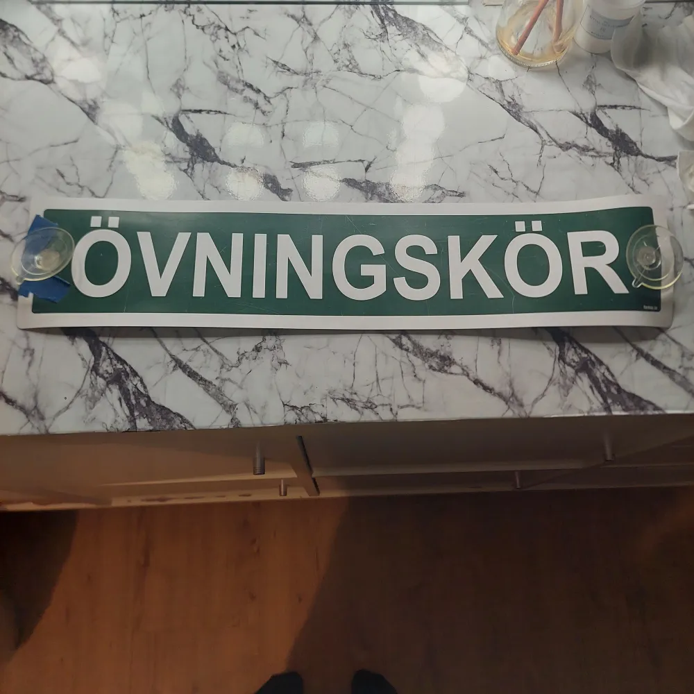 En övningskörnings skylt, sätts fast på insidan av bilrutan med hjälp av sugproppar. Vänstra sidan av skylten har gått sönder vid sugproppen och där av tejpen. . Övrigt.