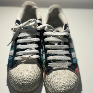 Adidas Superstar med blommigt mönster - Säljer ett par coola Adidas Superstar sneakers med ett unikt blommigt mönster. Skorna har den klassiska vita tåhättan och vita ränder på sidan. Perfekta för att ge din outfit en färgglad touch! De är i bra skick och redo för nya äventyr.
