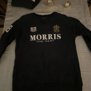 Morris tröja  - Säljer en snygg svart tröja från Morris med tryck på bröstet och en cool patch på ärmen. Perfekt för en stilren look, oavsett om du ska på fest eller bara hänga med vänner. Tröjan har långa ärmar och är i bra skick. Passar perfekt till jeans eller chinos!