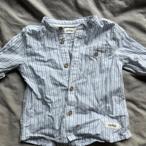 Randig skjorta från Newbie - Säljer en söt randig skjorta från Newbie i storlek 80 för barn 12-18 månader. Skjortan är ljusblå och vit med bruna knappar.