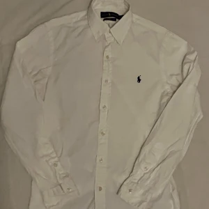 Vit skjorta från Ralph Lauren - Säljer en stilren vit skjorta från Ralph Lauren i storlek S. Skjortan är slim fit och gjord av lätt bomullstwill, perfekt för både vardag och fest. Den har långa ärmar och klassiska knappar framtill. En liten broderad logga på bröstet ger en extra touch av stil. Passar perfekt till både jeans och kostymbyxor!