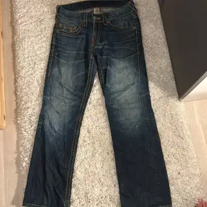 Säljer ett par snygga True Religion jeans i blå denim med coola broderade detaljer på bakfickorna. De har en klassisk femficksdesign och en bekväm passform. Perfekta för en avslappnad stil!