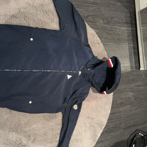 Mörkblå jacka från Moncler - Säljer en snygg mörkblå jacka från Moncler i storlek L. Jackan har en dragkedja framtill och en huva med röd och vit randig detalj. Perfekt för höst och vår, med en stilren look och Moncler-logga på ärmen. Passar både till vardags och lite finare tillfällen. Helt oanvänd och scannern på moncler logan funkar utmärkt 
