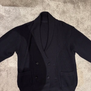 Mörkblå ribbad kofta - Säljer en snygg blå ribbad kofta med dubbelknäppning och stora fickor fram. Perfekt för höst och vinter, den är både stilren och varm. Koftan har långa ärmar och en skön passform som funkar till både vardag och fest. Passar perfekt över en t-shirt eller skjorta!