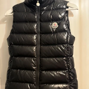 Svart dunväst från Moncler strl 152 eller 12år - Säljer en snygg svart dunväst från Moncler i mycket bra skick. Västen har en glansig yta och en dragkedja framtill. Perfekt när du vill hålla dig varm och stilren. Moncler-loggan är broderad på bröstet. Passar både till vardags och lite finare tillfällen.