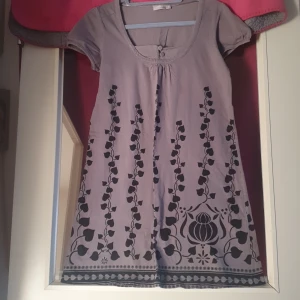 Ljuslila klänning med blommönster - Söt grå klänning med svart blommönster. Den har korta ärmar och en rund halsringning med en liten knappdetalj framtill. Perfekt för vår och sommar, med en avslappnad passform som gör den bekväm att bära hela dagen.