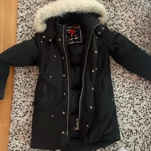 Svart vinterjacka från Moose Knuckles - Säljer en snygg svart vinterjacka från Moose Knuckles med pälskantad huva. Jackan har dragkedja och knappar framtill samt coola metallloggor på ärmarna. Perfekt för kalla vinterdagar! 🖤