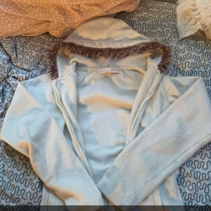 Vit fleecejacka med pälskant - Mysig vit fleecejacka med en snygg pälskant runt huvan. Perfekt för kyliga dagar när du vill hålla dig varm och bekväm. Jackan har långa ärmar och en dragkedja framtill. Passar bra till både vardag och utflykter. Dragkedjan funkar nt