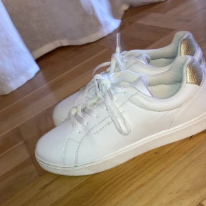Vita sneakers med gulddetaljer från Tommy Hilfiger - Säljer ett par snygga vita sneakers från Tommy Hilfiger i nytt skick. De har gulddetaljer på hälen och klassisk snörning. Perfekta för både vardag och festliga tillfällen. Storlek dam 41