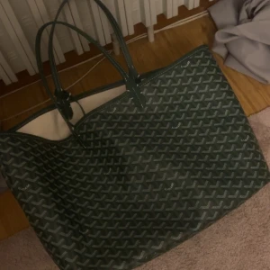 Grön väska - Säljer en snygg grön shoppingväska med ett klassiskt mönster. Väskan är rymlig och perfekt för shoppingturer eller som en vardagsväska. Den har två handtag och en liten innerficka för extra förvaring. Materialet verkar vara av syntet och insidan är ljus och rymlig.