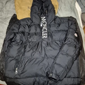Moncler maury - Säljer en snygg svart pufferjacka från Moncler i mycket bra skick. Jackan har en stor logga på framsidan och en praktisk dragkedja. Perfekt för kalla vinterdagar med sin varma och vadderade design. Den har också en cool etikett med serietidningsmotiv på insidan.
