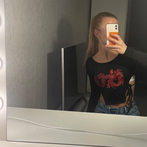 Svart långärmad topp med röd drake - Säljer en cool svart långärmad topp med en röd drake på framsidan. Toppen är croppad och har snörning i sidorna för en extra edgy look. Perfekt för en utekväll eller fest! 