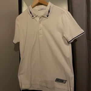 Exklusiv Givenchy Piké - Höj din avslappnade stil med denna exceptionella vita Givenchy herr Pikén i storlek M, oanvänd i splitternytt skick. Pikén är tillverkad i Italien av 100% bomull. Perfekt för en avslappnad men ändå sofistikerad look. Denna polotröja har en klassisk stängning med tre knappar och korta ärmar med en ribbad profil. Äkthetsintyg medföljer, inköpt för 7000kr, säljes för 3199kr.🤝🏻