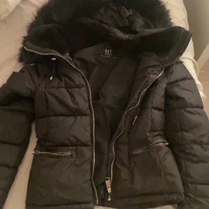 Svart pufferjacka från Zara - Säljer en snygg svart pufferjacka från Zara TRF Collection. Jackan har en mysig huva med fuskpäls och praktiska dragkedjor framtill och på fickorna. Perfekt för kalla vinterdagar! 🖤